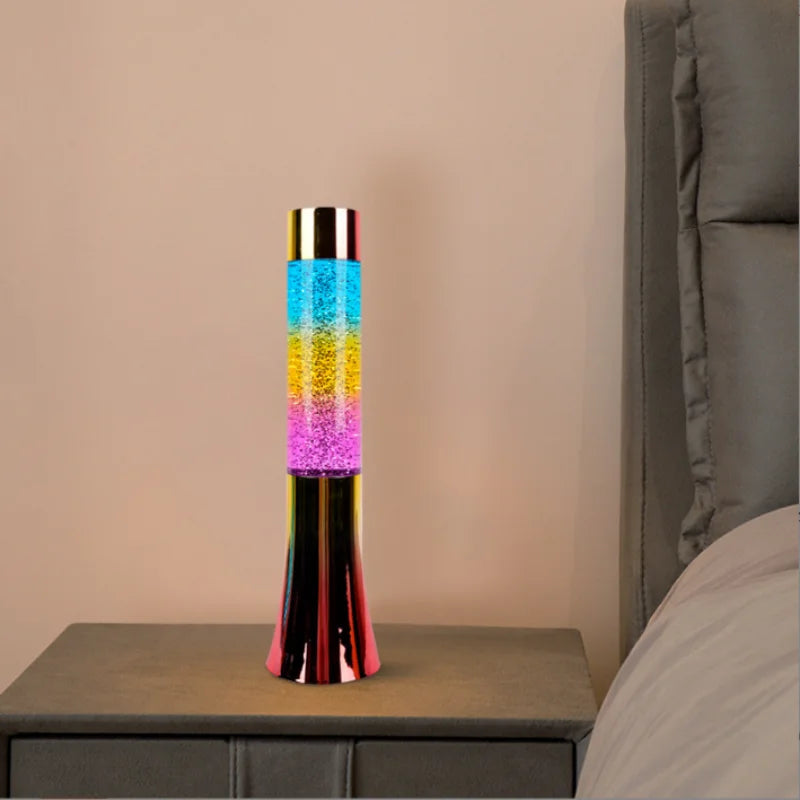 A Color-Changing LED Glitter Lava Lamp, 13 inches tall with layers of blue, yellow, orange, and purple gel, sits on a bedside table by a gray headboard. Its metallic base beautifully reflects the lighting.