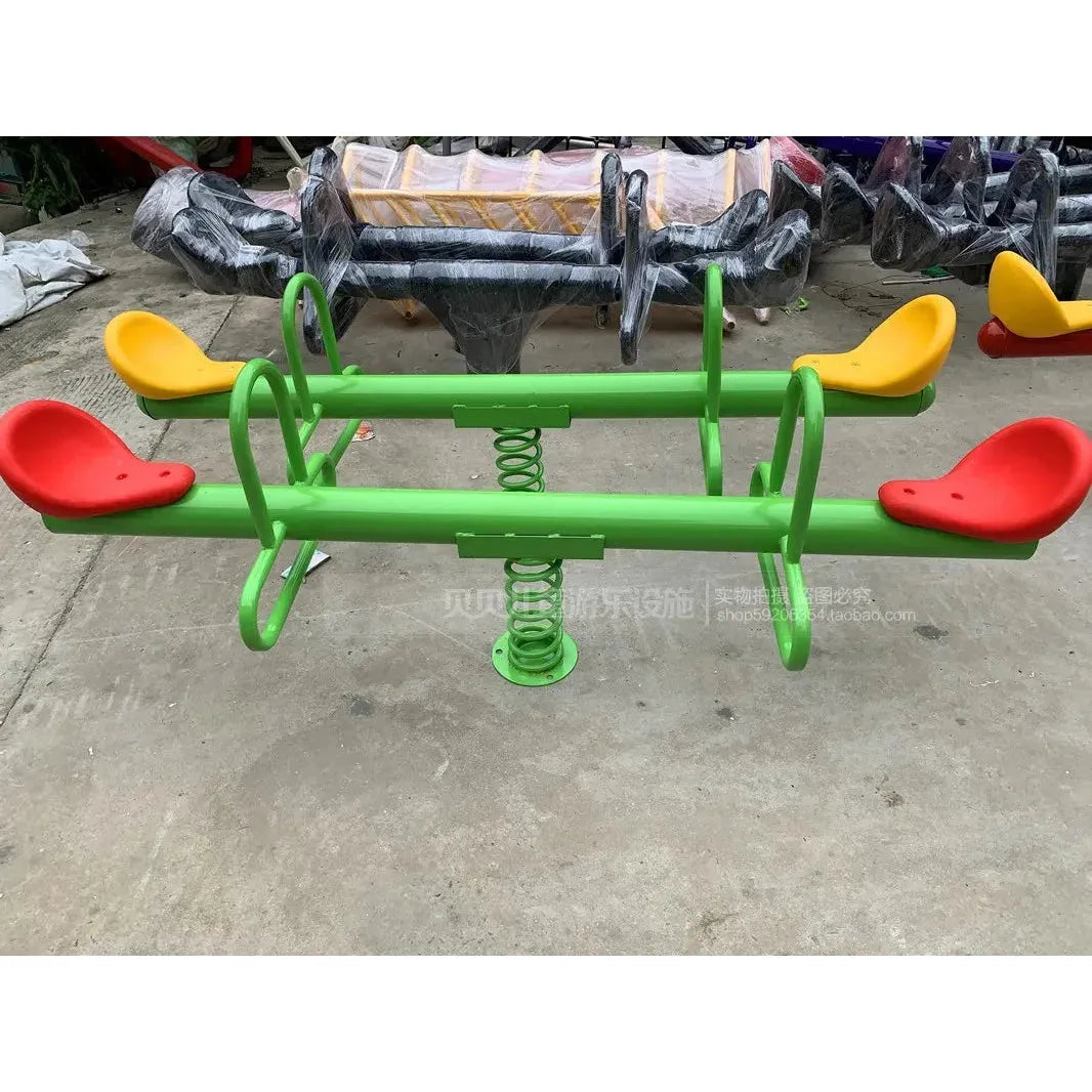 A green metal frame swing, ideal for childrens sensory integration, with red and yellow seats on coil springs is displayed in a playground. The outdoor steel climbing frame sits on concrete, with bags of additional equipment visible in the background.