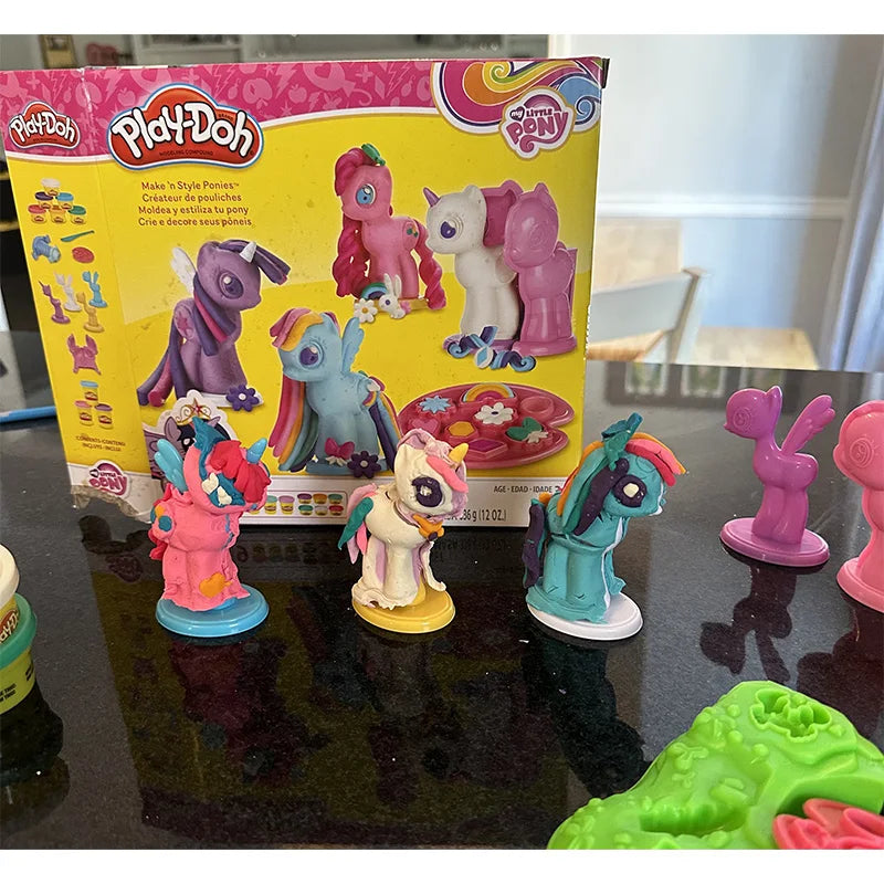 Colorful My Little Pony Play-Doh models and molds are arranged on a dark surface, with the Play-Doh box in the background featuring assembly instructions and images of vibrant, completed ponies.