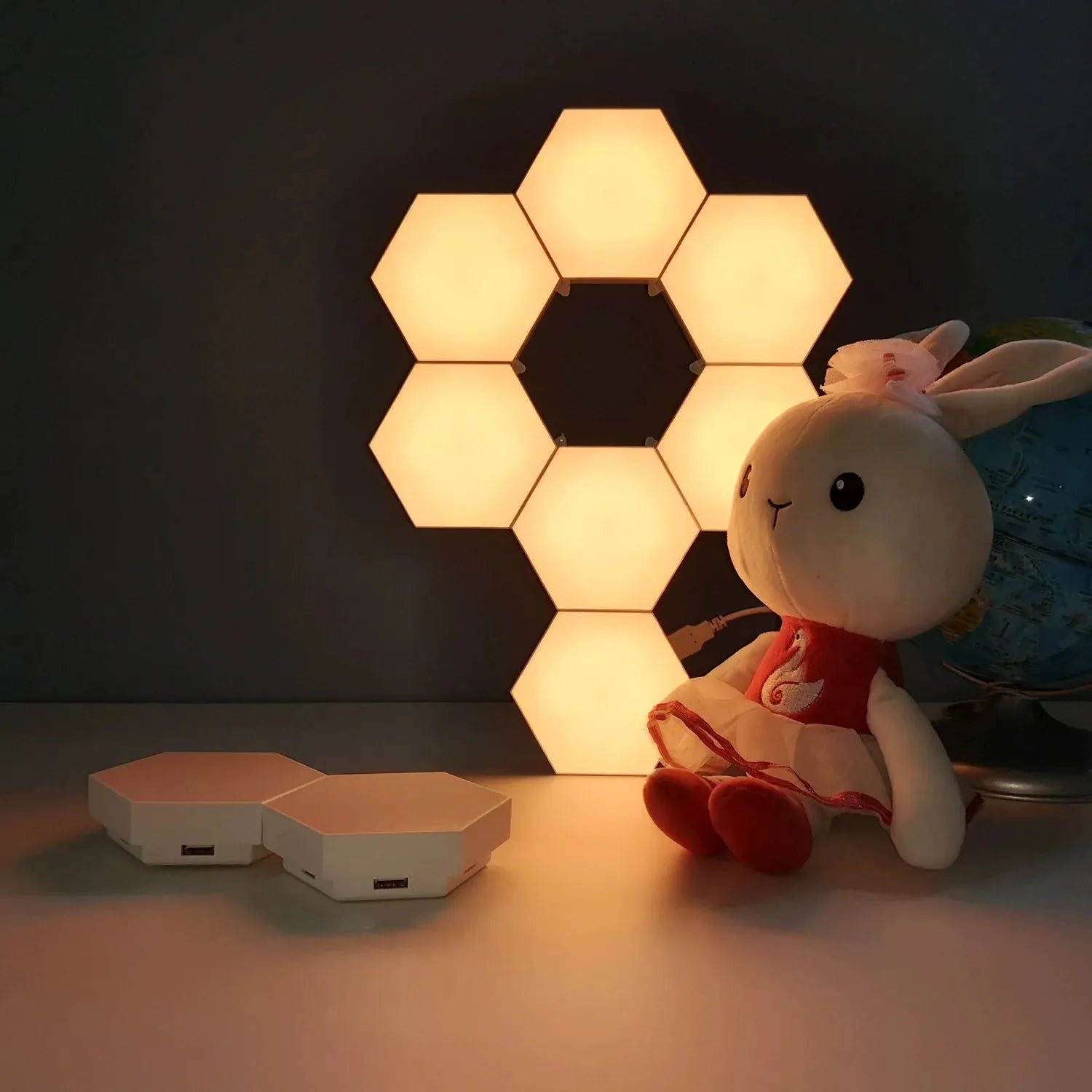 Hexagonal LED Modular Night Light Panels for Sensory Rooms glow warmly on a dark wall, while a plush white rabbit toy in a red dress sits beside them. The calming lamps offer soothing light, perfect for autism or sensory spaces. A globe rests nearby.