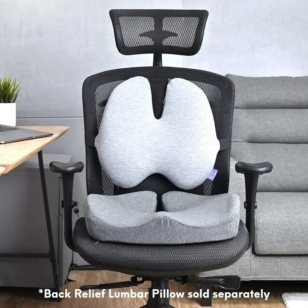 An ergonomic office chair with a Cushion Lab Patented Pressure Relief Seat Cushion for long sitting hours and a gray memory foam lumbar pillow is in front of a desk. The chair features a mesh back, headrest, and adjustable armrests. Text below reads *Back Relief Lumbar Pillow sold separately.