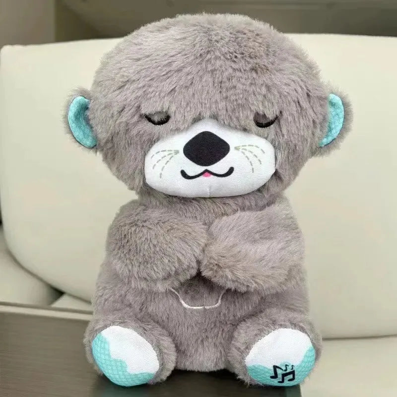 A silver plush otter sits serenely with a peaceful expression, eyes closed. It features turquoise ears and paws, plus a musical note on one paw, signaling its function as a soothing musical toy. Perfect for calming sensory experiences, it rests peacefully on the beige sofa.
