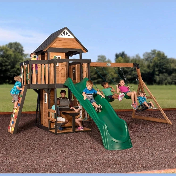 Children play on the Playhouse Backyard Discovery Canyon Creek Cedar Swing Set, which features a green slide, swings, climbing wall, and playhouse. Some swing or climb while others sit at a table beneath this outdoor playground set. Trees and grass surround them.