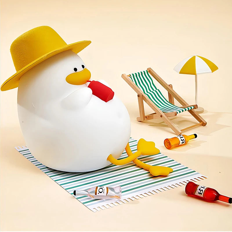 A cute, round duck in a yellow hat sits on a striped mat with a red drink and the Cute Cartoon Duck Rechargeable LED Night Light nearby. A small striped deck chair, beach umbrella, three small bottles, and a red ketchup bottle complete the scene against the light sandy background.