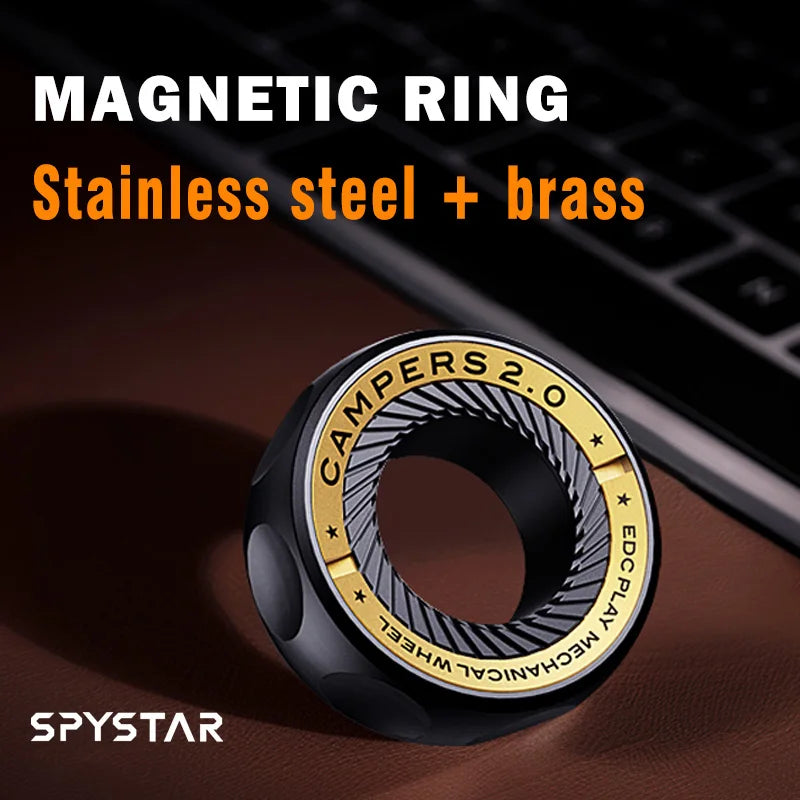 A black and gold Spystar Mechanic Ring Fidget Spinner Fingertip Gyro rests on a brown surface near a laptop. This magnetic metal adult anti-stress toy doubles as a stylish EDC ring. Text reads: MAGNETIC RING Stainless steel + brass.