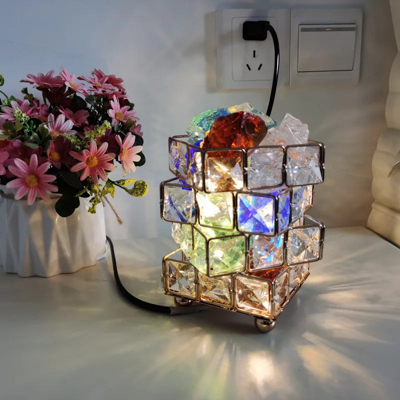 The Himalayan Salt Crystal LED Lamp, with colorful cube glass pieces, creates a warm table ambiance. Nearby, a vase holds pink and white daisies. A power outlet and USB plug add convenience in the background.