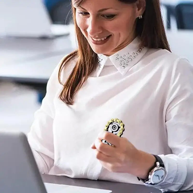 A woman wearing a white shirt with decorative collar accents sits at a table using her laptop. Shes holding a Sprockets Flywheel Fingertip Gyro Fidget Spinner, smiling as she explores new dropshipping opportunities on the screen.