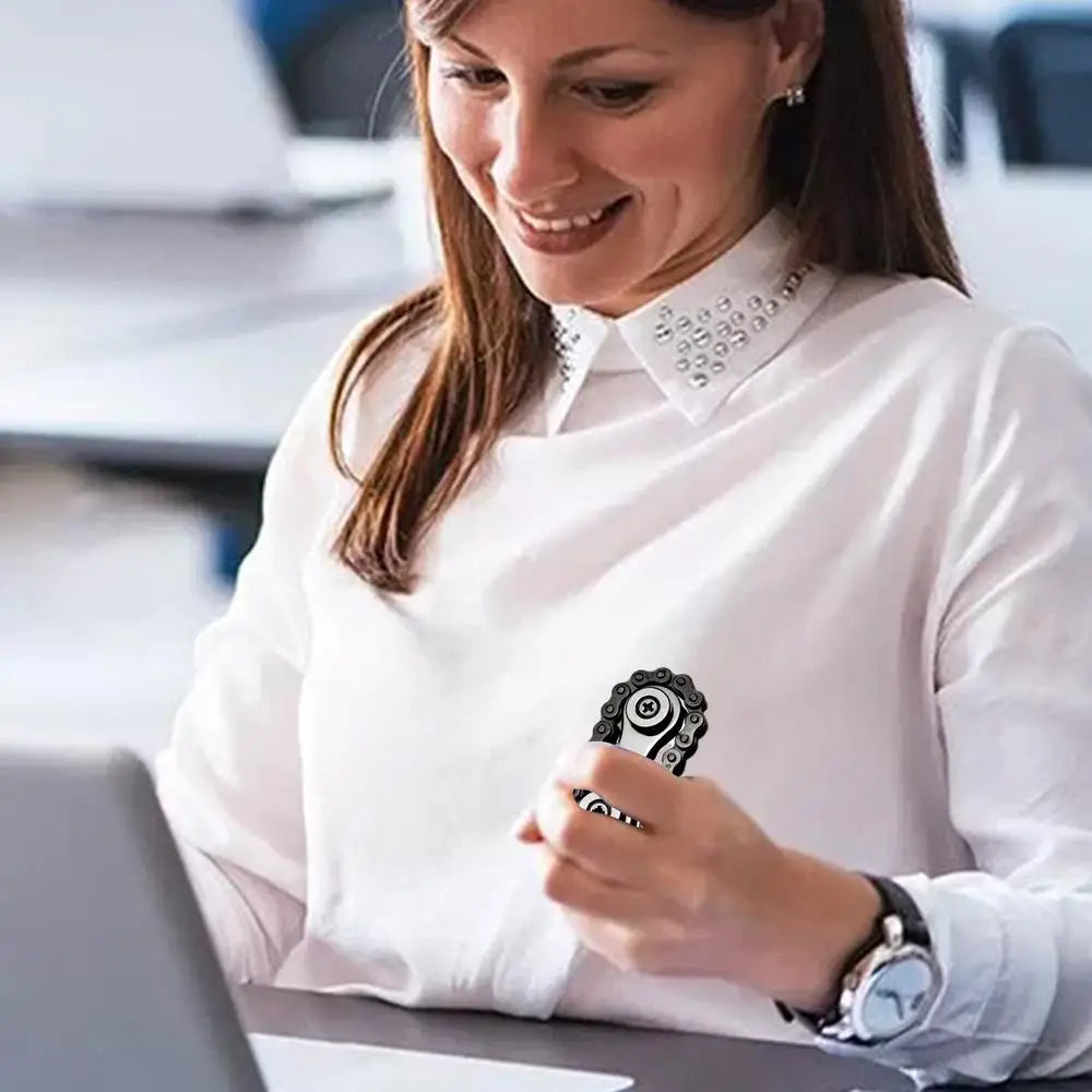 A smiling woman in a white blouse sits at a desk, holding a sleek Sprockets Flywheel Fingertip Gyro Fidget Spinner. Positioned before her laptop, she’s immersed in her dropshipping business within the brightly lit office setting.