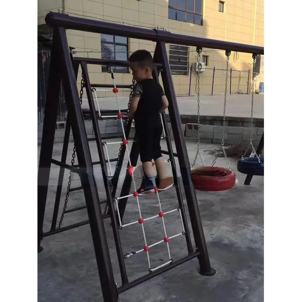 A young boy dressed in black climbs a rope ladder on the Outdoor Steel Climbing Frame Swing for Childrens Sensory Integration Training. Behind him, swings sway under clear skies, while a red and black tire swing stands out among the playground equipment.