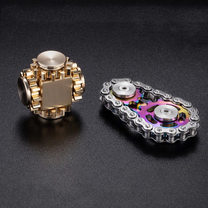 Two metallic fidget toys sit on a dark surface: a cube-shaped magnetic slider with gear-like edges and an iridescent bike chain metal fidget spinner, both ideal for stress relief, available as the Bike Chain Metal Fidget Spinner with Magnetic Slider - 9 Gear Linkage Stress Relief Toy.