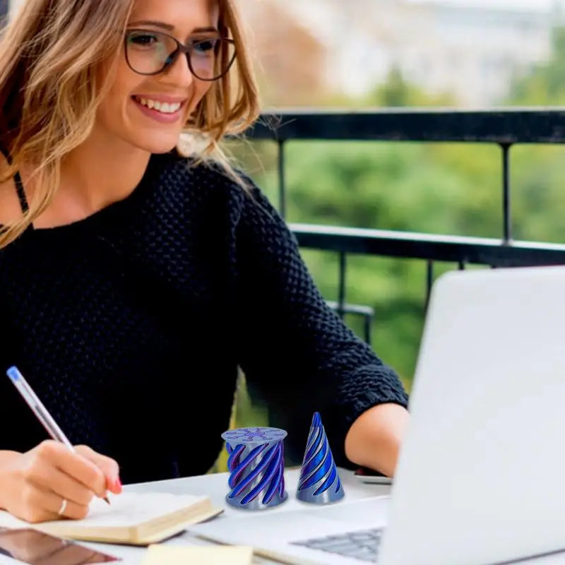 A woman with glasses smiles while working on a laptop outdoors. Beside her, two Impossible Cone Sensory Fidget Toys add creative flair to her workspace as she writes in a notebook.