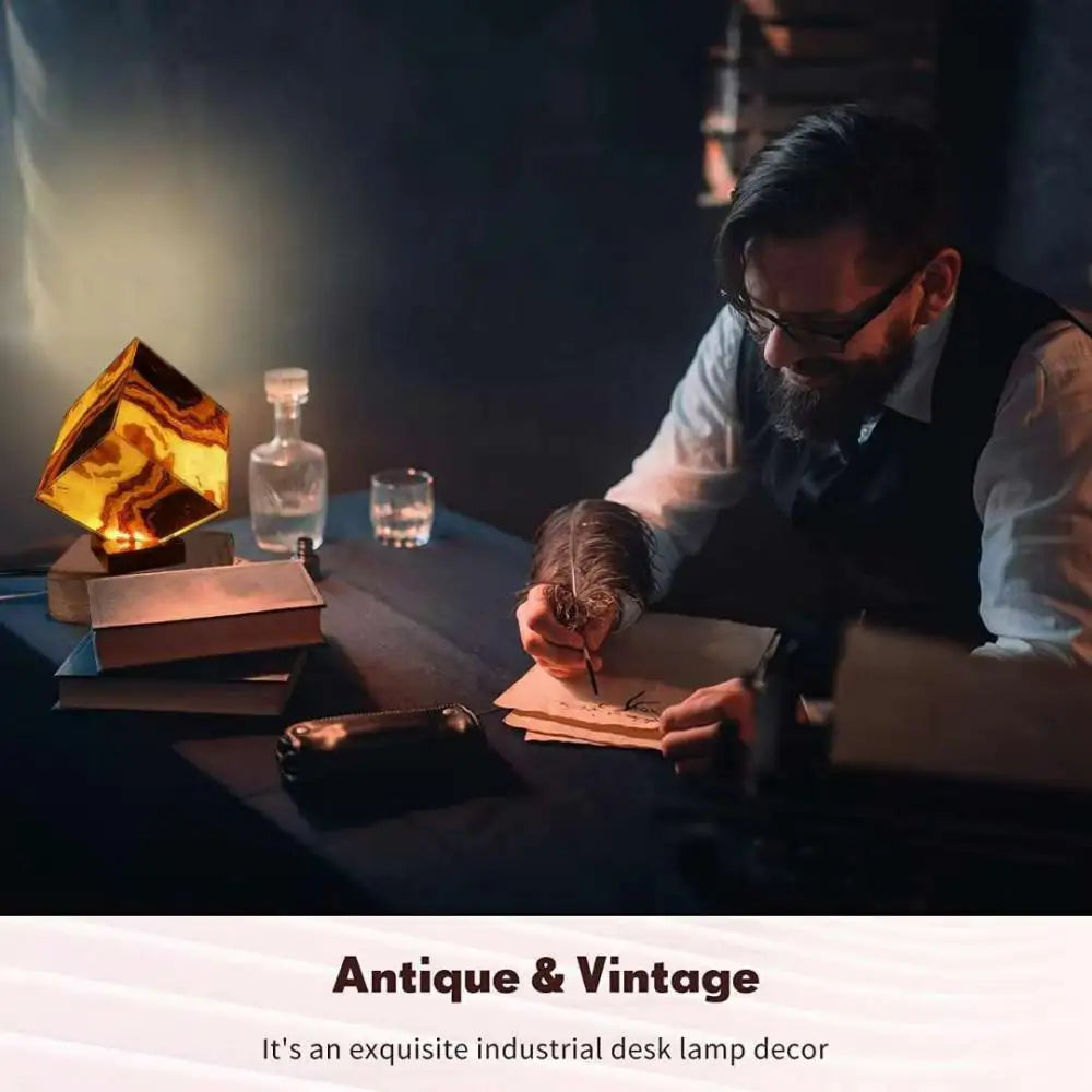 A bearded man with glasses writes with a quill at a dimly lit desk, where an amber-hued geometric lamp, glass bottles, and books adorn the table. The caption reads, Elegant Agate Crystal Table Lamp: Stylish Bedroom & Living Room Décor Light.
