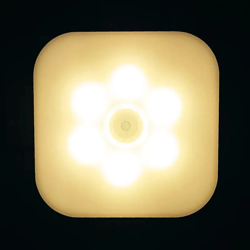 A close-up of the Dimmable Motion Sensor LED Night Light shows its square, rounded-corner design emitting a warm, yellow glow. The central bulb and five smaller lights form a flower pattern on a dark background, offering an energy-efficient lighting solution for any room.