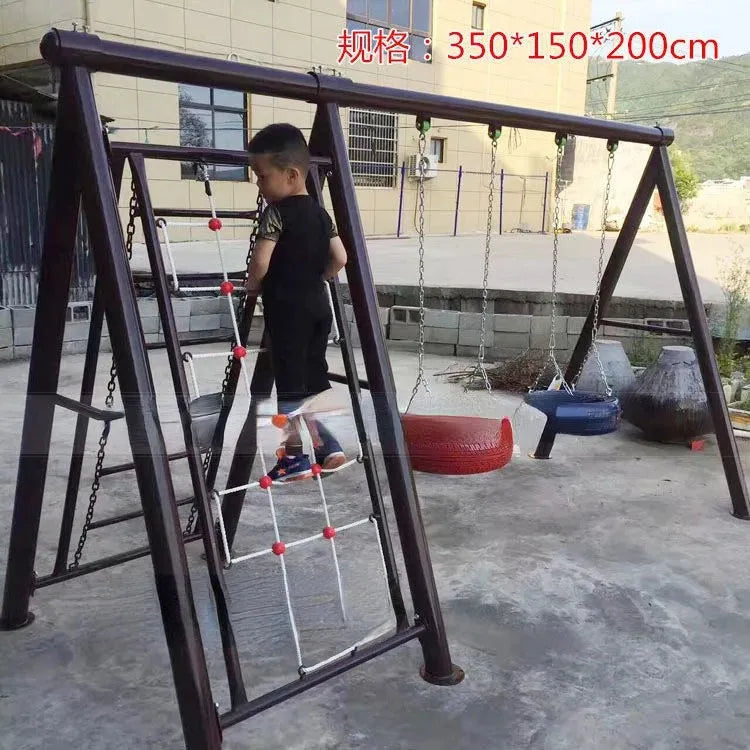 A child in a black outfit ascends an Outdoor Steel Climbing Frame Swing for Childrens Sensory Integration Training, featuring a ladder and two swings, one crafted from a red tire. Situated on concrete beside a building and scenic hills, this play structure enhances sensory development.