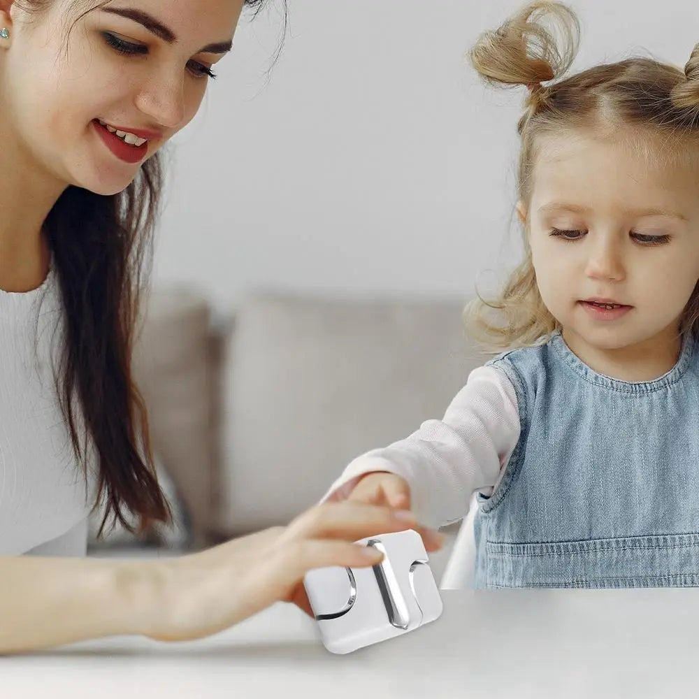 A woman and a young girl sit at a table. The girl, in a blue dress, reaches toward a silver Infinity Gear Cube Spinner Antistress Toy held by the smiling woman.