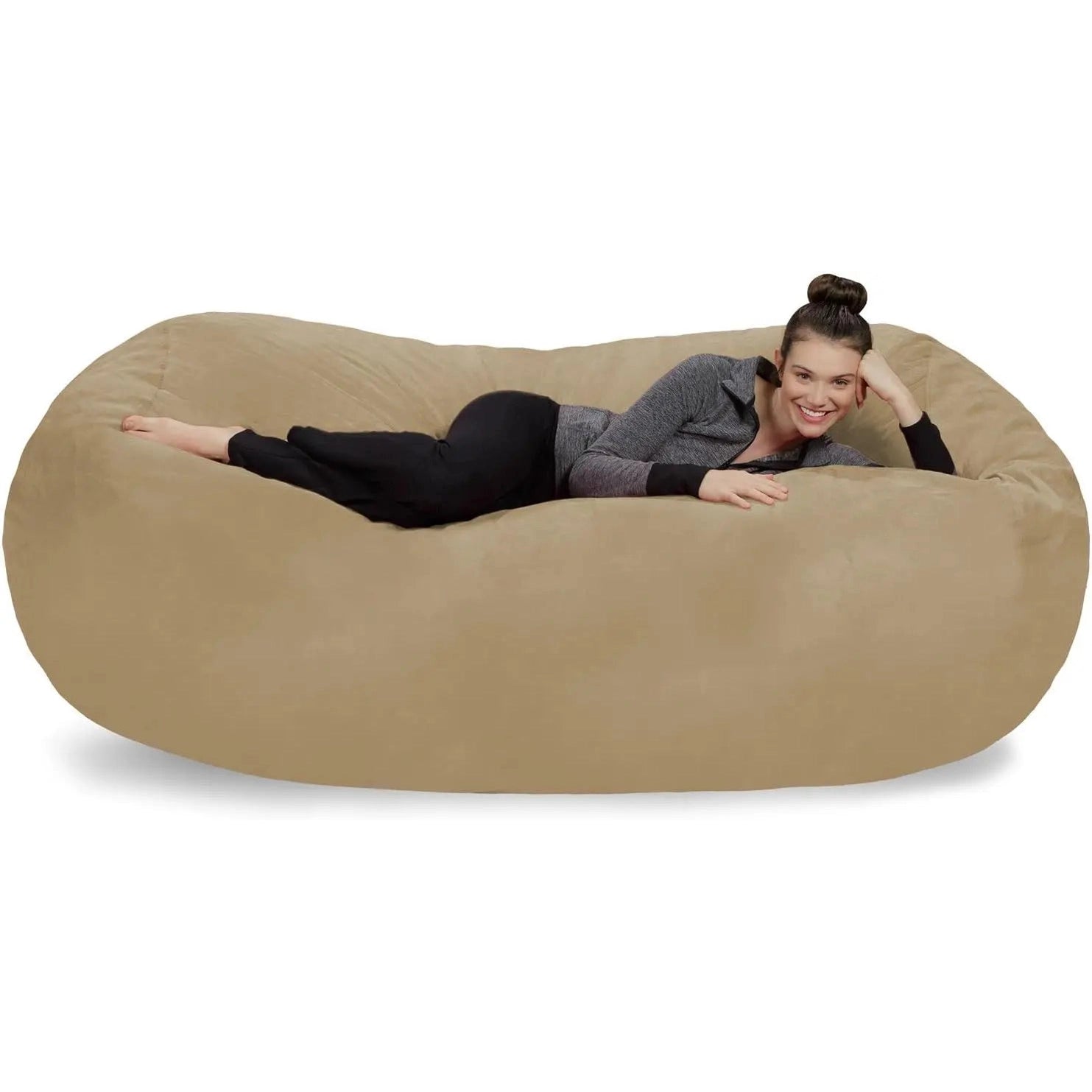 A woman with dark hair in a bun, wearing a gray sweater and black pants, is lying and smiling on a camel Bean Bag Lounger - Plush Sofas with Soft Microsuede Cover against a white background.
