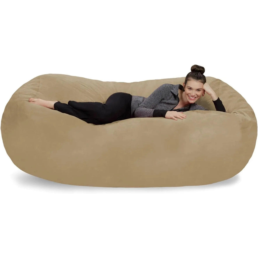 A woman with dark hair in a bun, wearing a gray sweater and black pants, is lying and smiling on a camel Bean Bag Lounger - Plush Sofas with Soft Microsuede Cover against a white background.