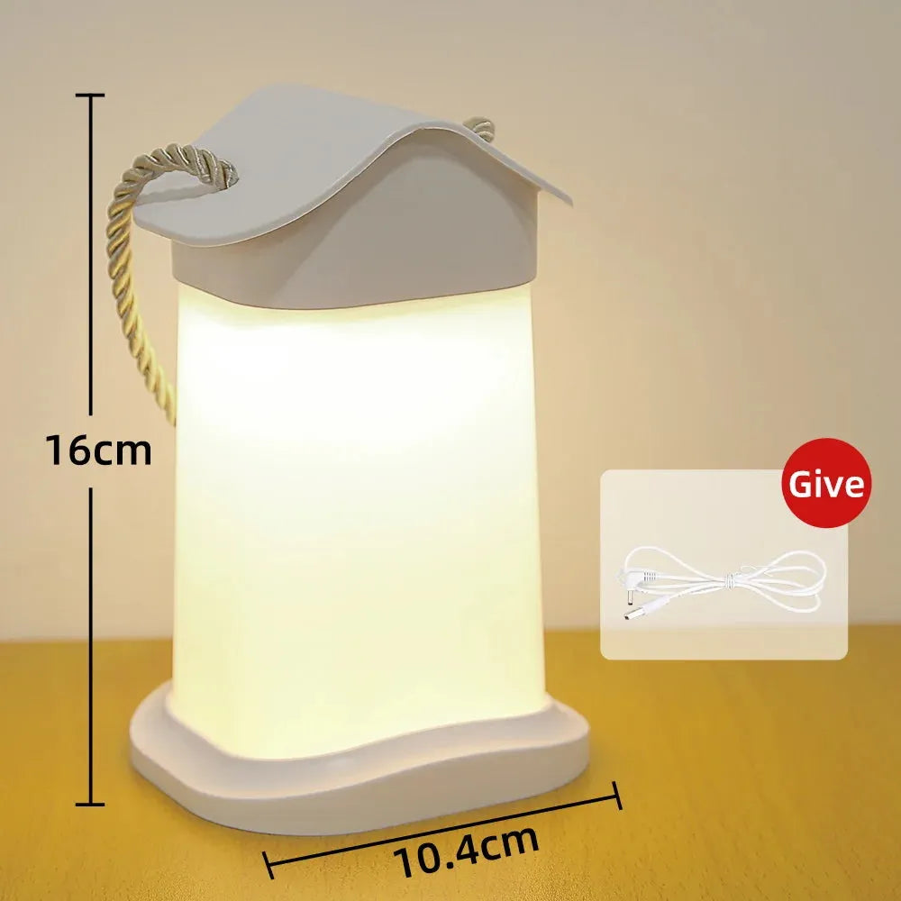 The Rechargeable Dimmable Touch LED Night Light is a white, lantern-shaped table lamp with a rope handle, glowing softly for your bedside. It measures 16 cm by 10.4 cm and comes with a charging cable labeled Give. Perfect as a Christmas gift for kids.