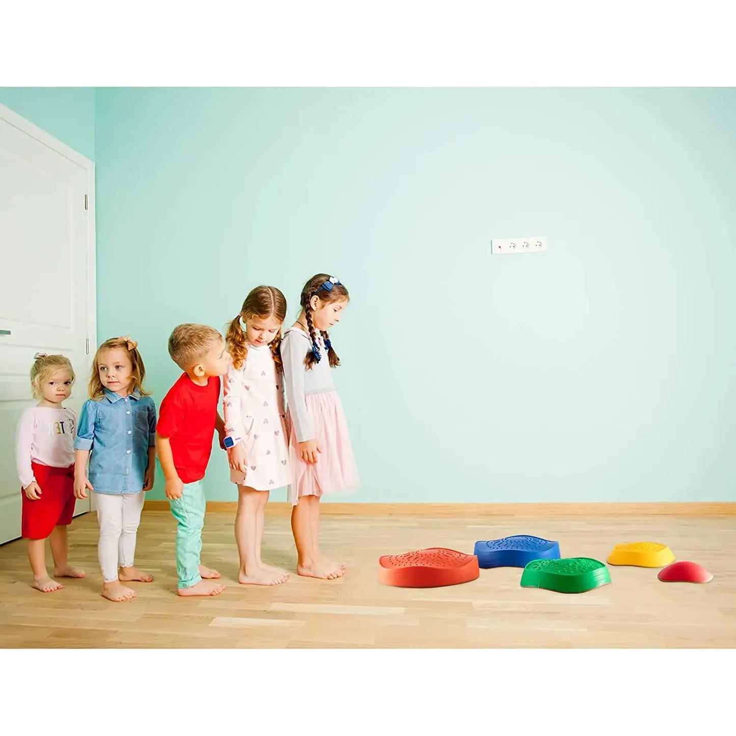 Five children stand indoors on a wooden floor, eagerly eyeing the vibrant Kids 5-Piece Outdoor Balance Stepping Stones Set for Coordination and Obstacle Play in red, green, blue, and yellow. The light blue walls and a white door are visible beside them.