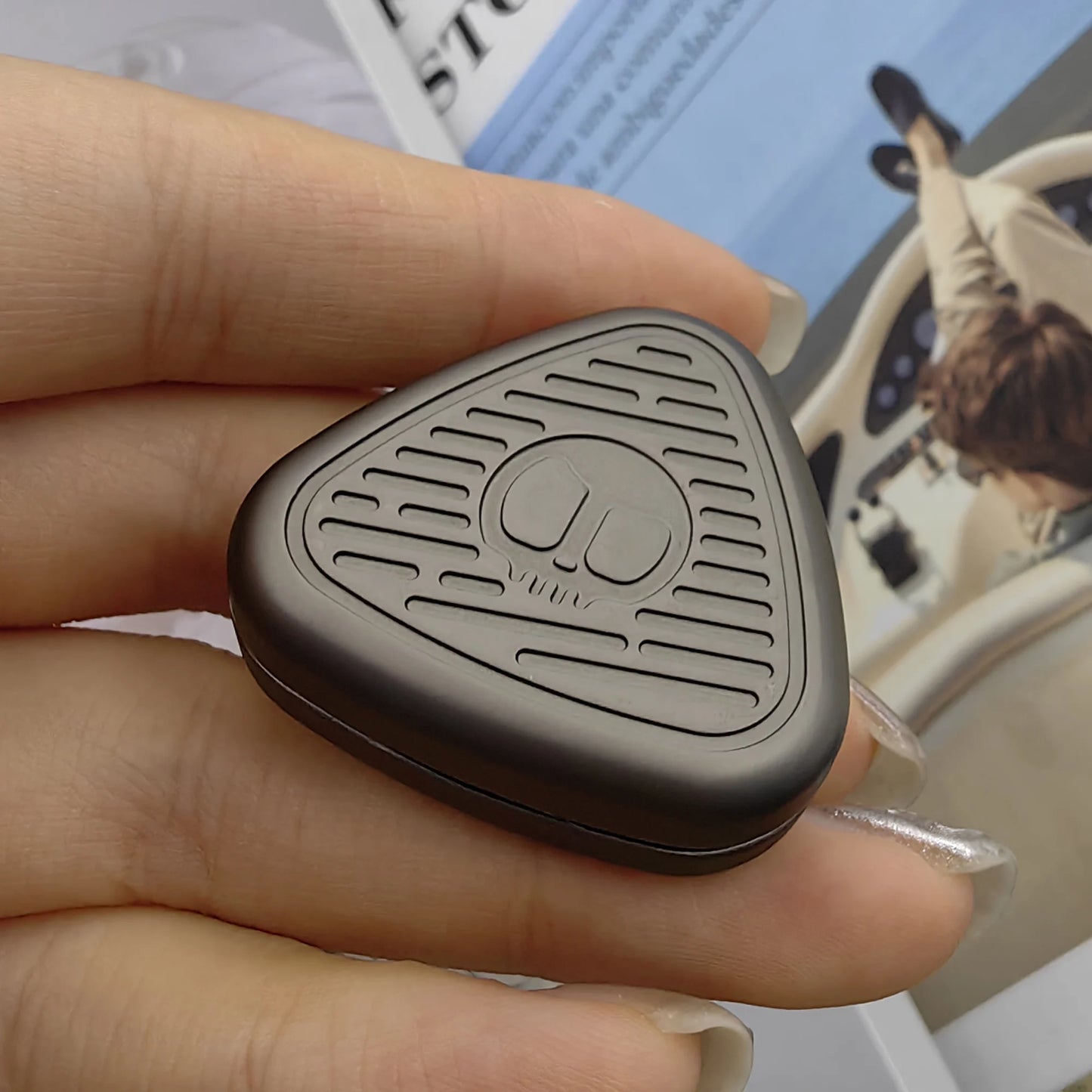 A hand with manicured nails holds a small, dark EDC Fingertip Toys Fidget Slider with a geometric metal grill design and embossed logo, against a blurred background featuring a magazine page showing someone at a steering wheel.