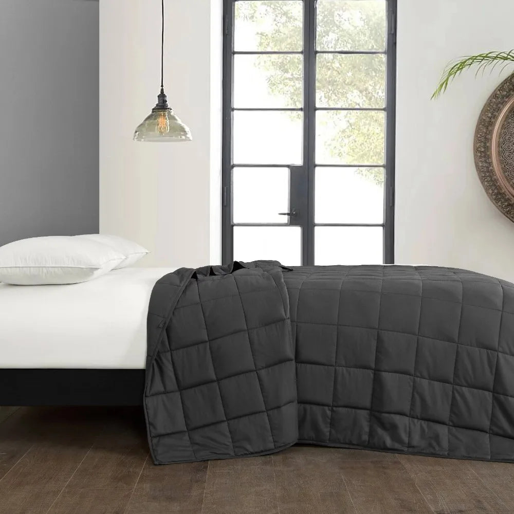 A modern bedroom features a bed with a dark gray Plush 20lb Soft Weighted Blanket, perfectly complemented by white pillows. In the background, a large black-framed window, decorative hanging light, and round wall mirror blend elegantly to highlight this US design.