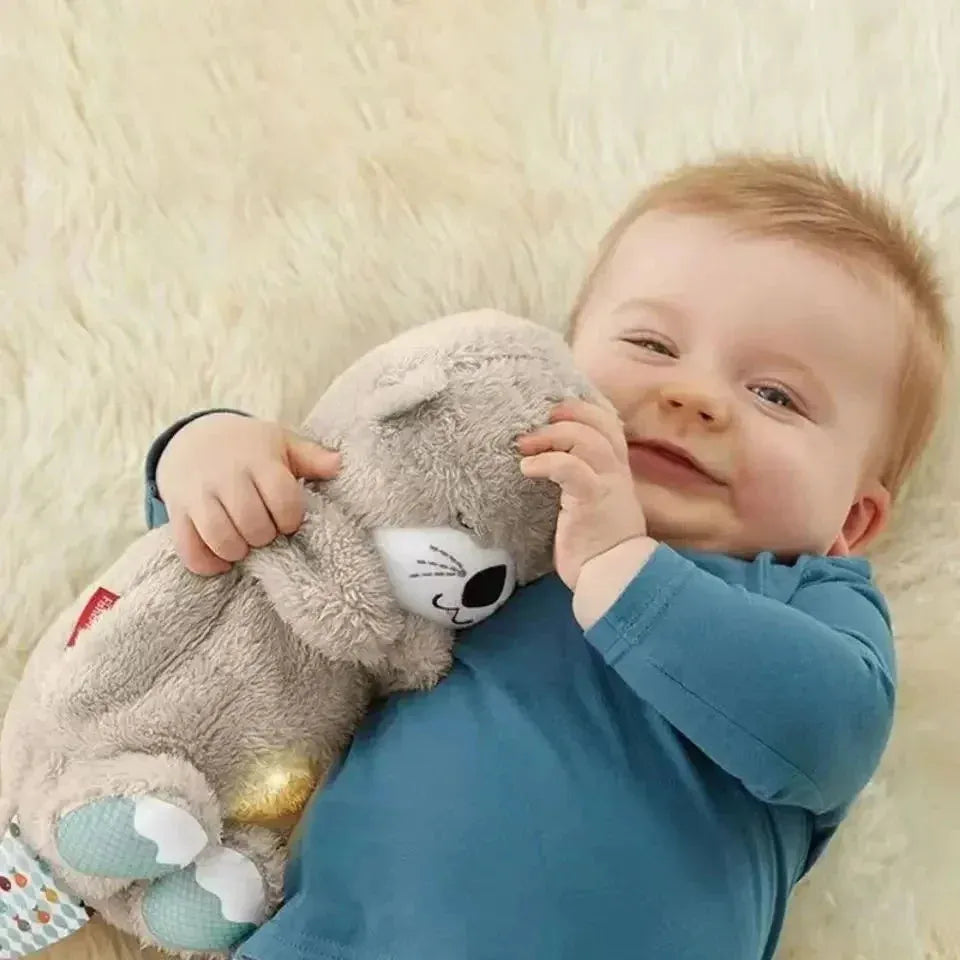 A smiling baby with light hair in a blue onesie lies on a cream blanket, playfully hugging the Soothing Otter Musical Plush Toy with Light and Sound for Newborns, enjoying its comforting sensory experience.