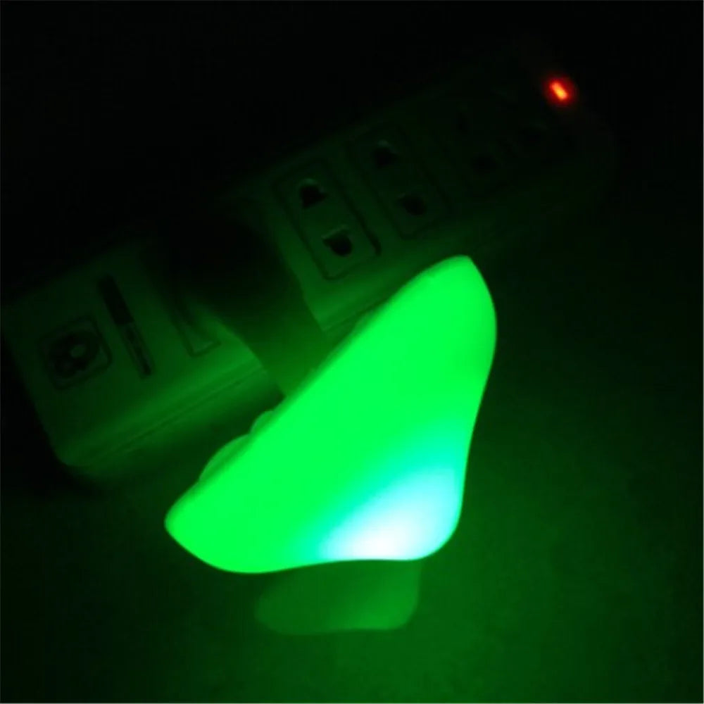 The Mushroom LED Night Light with Sensor Control, a warm white wall lamp, is plugged into a power strip in a dark scene. It emits a soft glow to illuminate the area while one red indicator light on the power strip is visible.