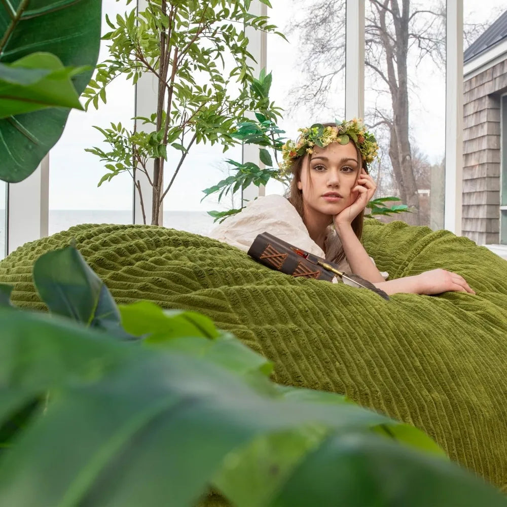 A woman in a floral crown and white dress lounges on a Fuf XXL Foam Filled Bean Bag Chair, Moss Green, 6 Feet, surrounded by lush indoor plants in a bright room with removable cover for easy care.