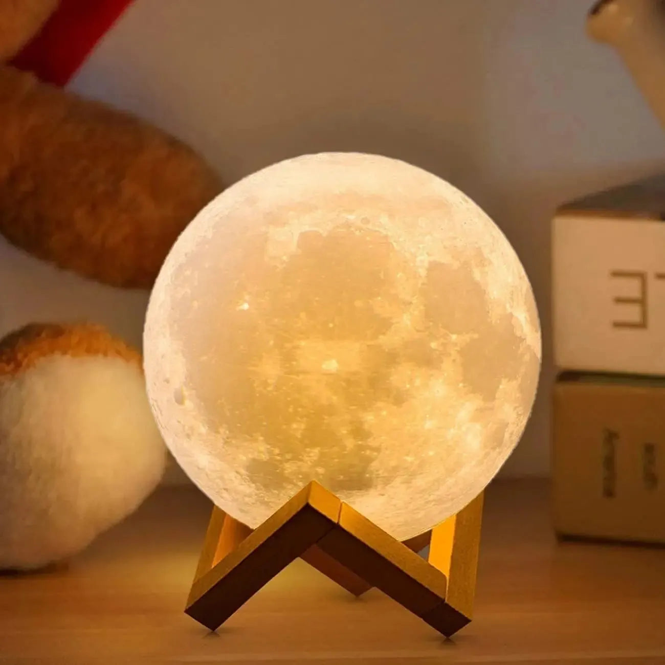 The Celestial 3D Moon Lamp LED Night Light, resembling the moon with its warm glow, beautifully rests on a wooden stand. On the left, a plush toy peeks out, while to the right, stacked books fade softly into the background.