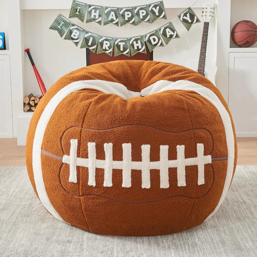The Sports Football Bean Bag Chair for Adults and Kids in Football color, crafted with ultra-soft sherpa plush, sits in a sports-themed living room decorated with a Happy Birthday banner, guitar, bat, basketball, and stacked firewood.