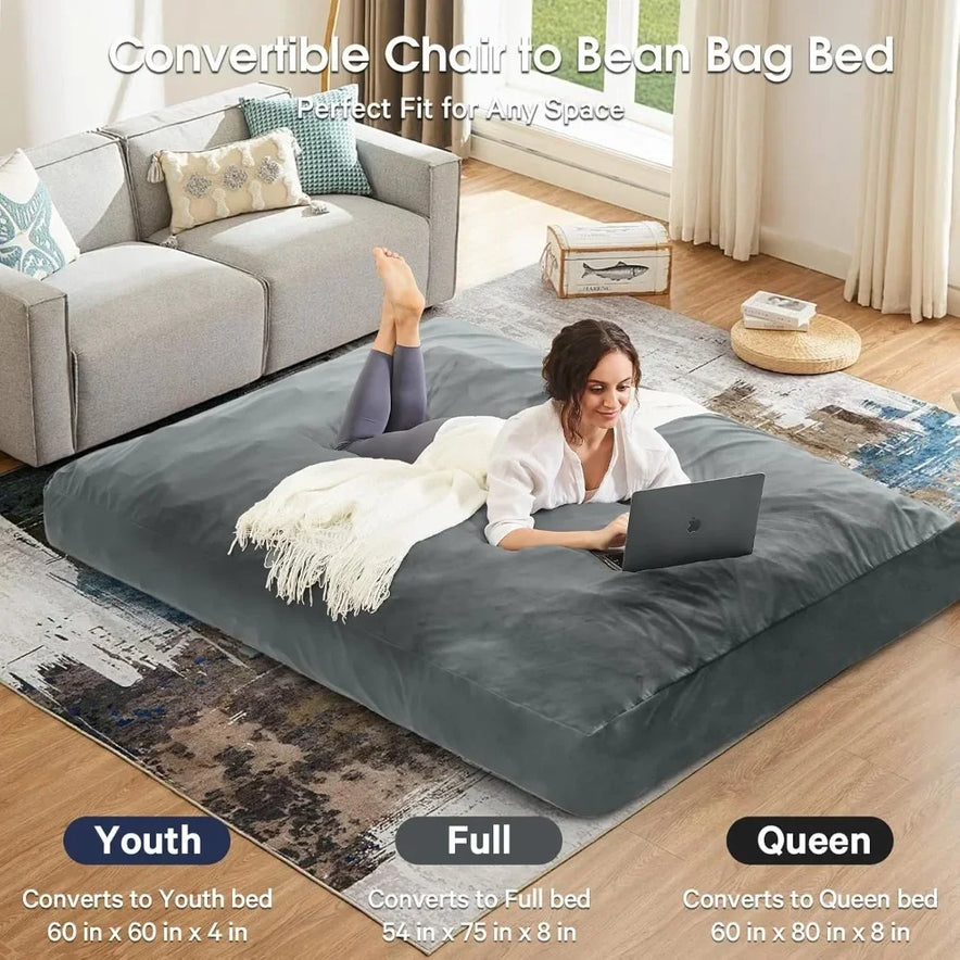 A woman lies on her stomach using a laptop on a Bean Bag Chairs Convertible Bean Bag Bed with a gray microfiber cover, shown in a living room. The bed comes in Youth, Full, and Queen sizes. A gray sofa and decor are visible in the background.