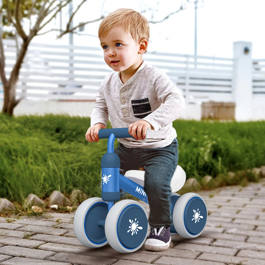 A young child in a gray shirt and jeans rides the Baby Balance Bike Ride On Toy for 1 Year Old Boys and Girls, an indoor anti-drop baby walker for ages 10 to 24 months, on a paved path with grass and a white fence in the background.