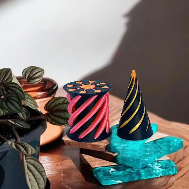 The Impossible Cone Sensory Fidget Toy—a 3D printed pyramid passthrough sculpture with a yellow spiral—rests on a wooden surface beside blue glass, plants, and a candle in warm office light.