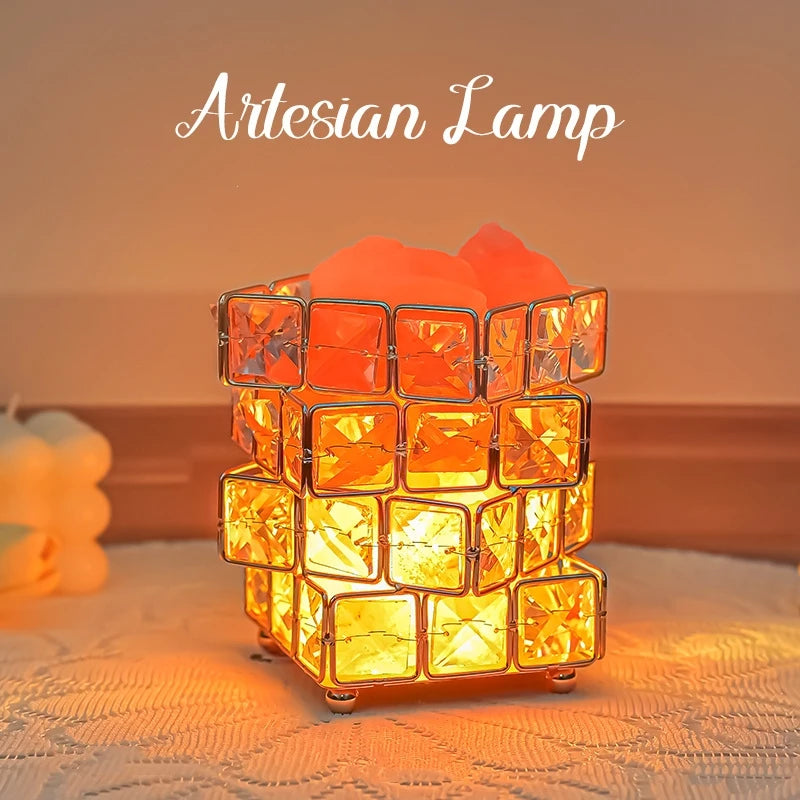 The Himalayan Salt Crystal LED Lamp, a USB night light for home décor and relaxation, emits a warm, ambient glow reminiscent of an Artesian lamp. It features a square design with bright gemstones in a metal grid, sitting on a textured surface with soft orange lighting in the background.