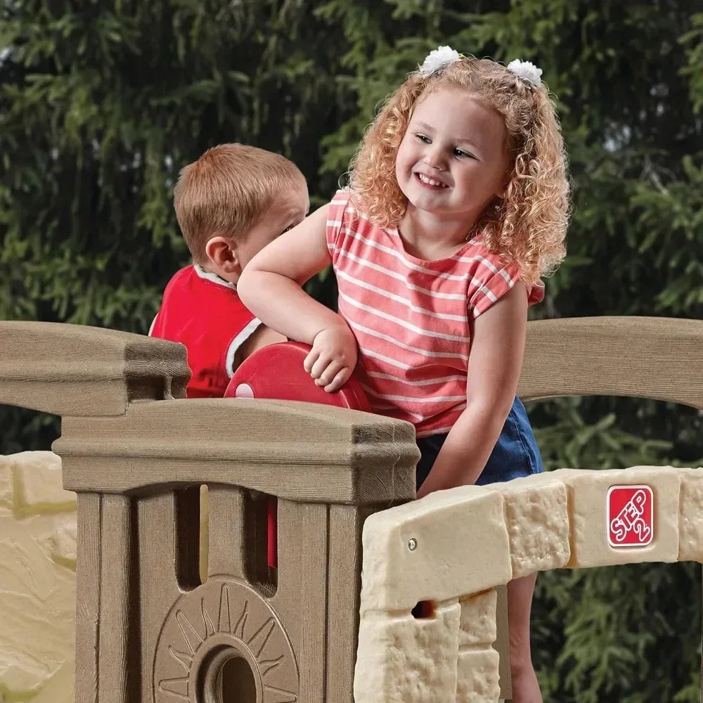 Two young children play outdoors on the Woodland Climber II Kids Playset for Ages 2 to 6 Years. A smiling girl with curly blonde hair wears a pink striped shirt, while the boy in red faces away. Trees surround the playset in the background.