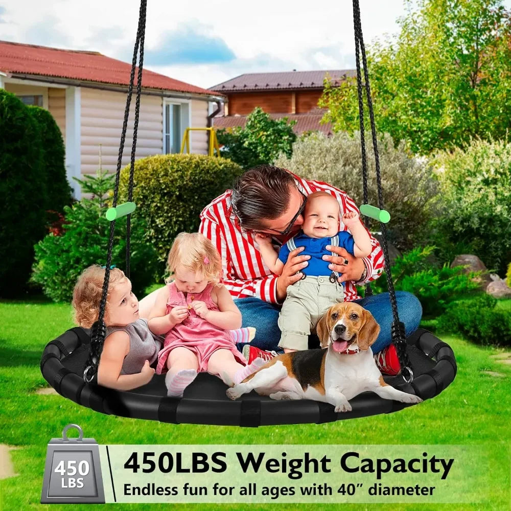 A man and three kids smile and play with a beagle on a Tree Swing with PP Mat for Kids and Adults, Black Outdoor Saucer Swing 40 Diameter, 450 lbs weight capacity. They sit together in a garden with a house in the background.