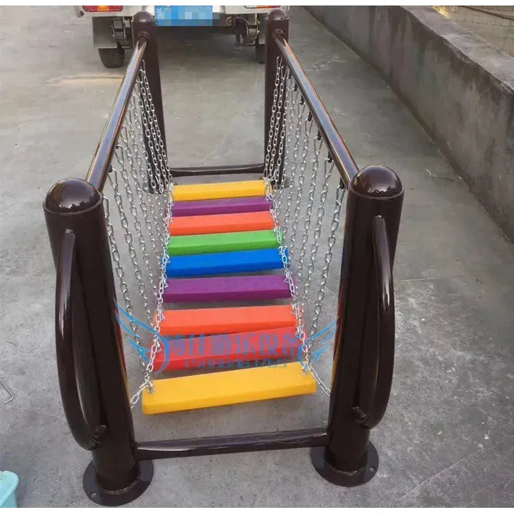 A unisex swinging bridge with rainbow wooden planks, suspended by chains and framed by dark brown metal railings, sits on concrete. It resembles a metal toy and is part of the outdoor toys Childrens swings Climbing nets Swinging bridges for ages 3-8, encouraging playful exploration.