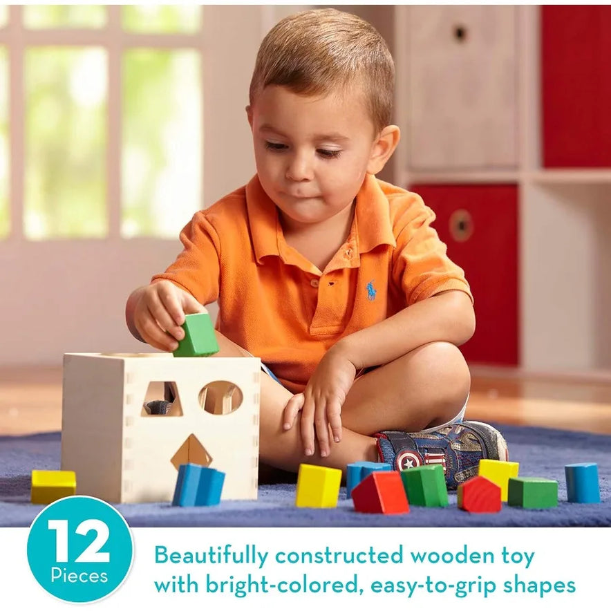 A young child in an orange shirt sits on the floor playing with the Melissa and Doug Geometric Stacker Wooden Educational Toy Shape Sorting Cube, featuring 12 colorful, easy-to-grip shapes. Blocks are scattered around. Educational fun for little learners!.