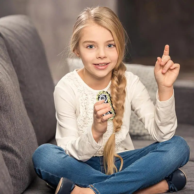 A young girl with long blonde hair sits cross-legged on a gray couch, wearing a white long-sleeve shirt and blue jeans. She holds the Sprockets Flywheel Fingertip Gyro Fidget Spinner in one hand and raises a finger with the other, smiling slightly.
