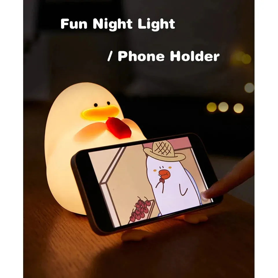 A cute, duck-shaped silicone night light holds a smartphone showing a cartoon penguin with a hat. Its LED glow adds charm, and the text above reads Cute Cartoon Duck Rechargeable LED Night Light – Perfect Birthday Gift.