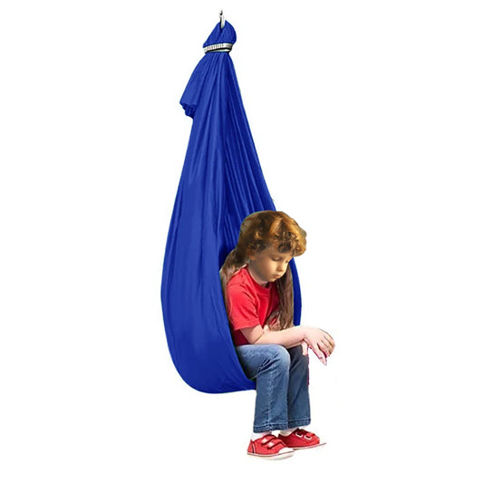 A child with curly hair, wearing a red shirt and jeans, sits in an Extra-Large Blue Sensory Swing Hammock for Kids and Adults with Special Needs (59 x 110). The swing offers a therapeutic experience. Relaxed with their hands on their knees in red sneakers, the background is plain white.