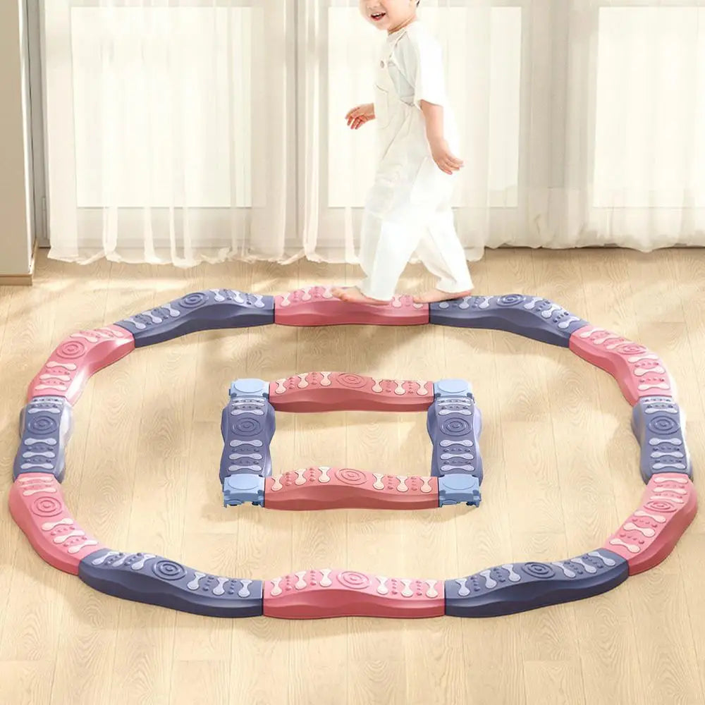 A child balances on the Sensory Balance Beam Training Board for Kids, arranged in circular and square patterns on a wooden floor. Pink, purple, and blue toys enhance motor skills as sunlight streams through large windows.