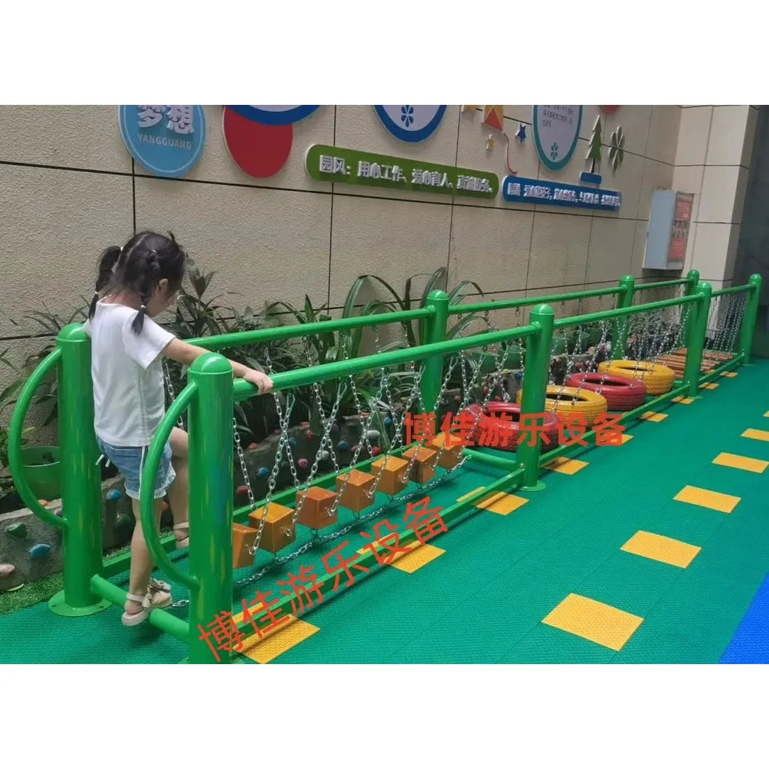 A child enjoys an outdoor playground structure featuring climbing nets, swinging bridges, and wooden steps suitable for ages 3-8. The floor is covered with interlocking green mats, with company signs visible on the wall in the background.