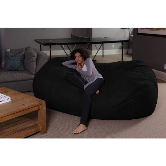 A woman relaxes thoughtfully on the Bean Bag Lounger - Plush Sofas with Soft Microsuede Cover in a living room with a wooden coffee table, gray couch, and ping pong table, resting her arm on the lounger.