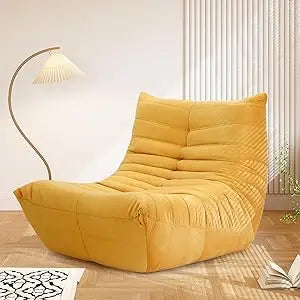 A yellow Fireside Chair Bean Bag for adults with plush memory foam and soft suede fabric rests on a wooden floor beside a curved floor lamp and open book, with vertical white blinds in the background.