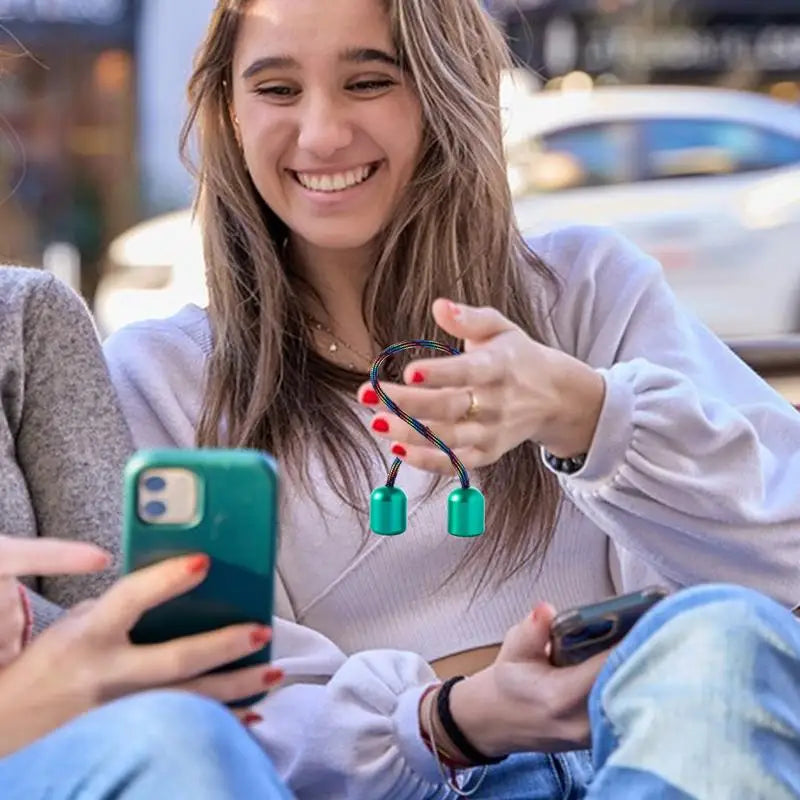 Two young women sit outdoors, smiling at a smartphone. One holds the Fidget Toy Worry Beads Finger Skill Anxiety Relief Kinetic Art for Men and Women, ideal for easing anxiety. Cars and buildings appear blurred in the background.