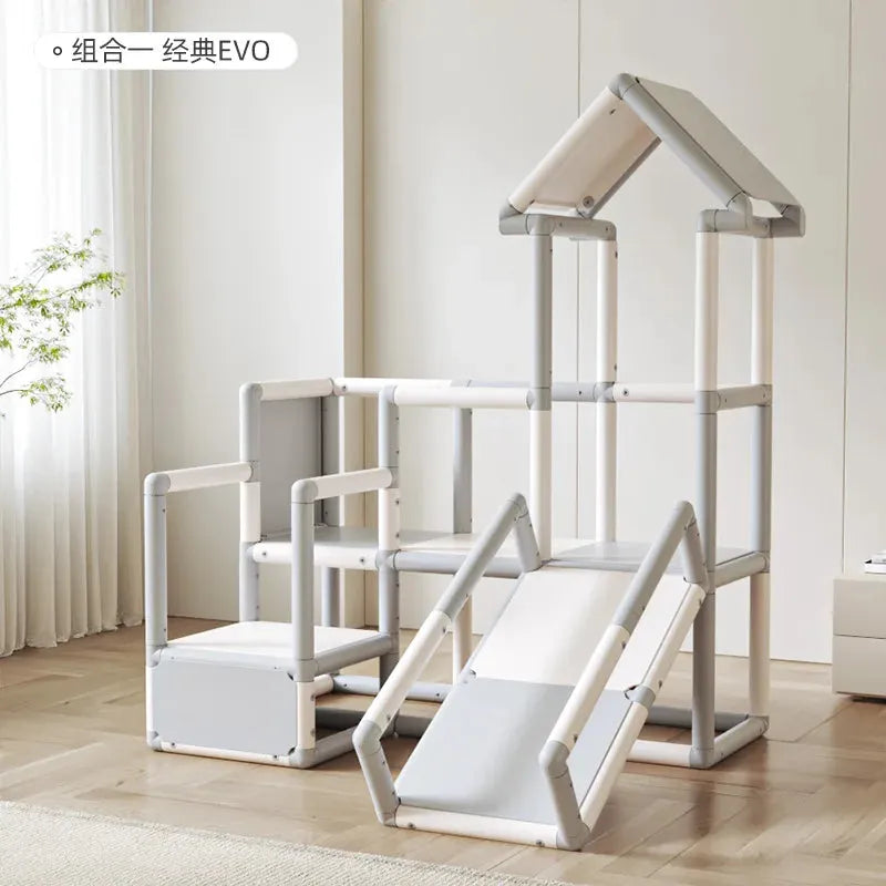 The Indoor Climbing Adventure Set for Kids includes a baby slide, climbing areas, and a top frame resembling a playhouse. Mainly white and light gray, it sits on a wooden floor in a bright room with a potted plant in the corner.