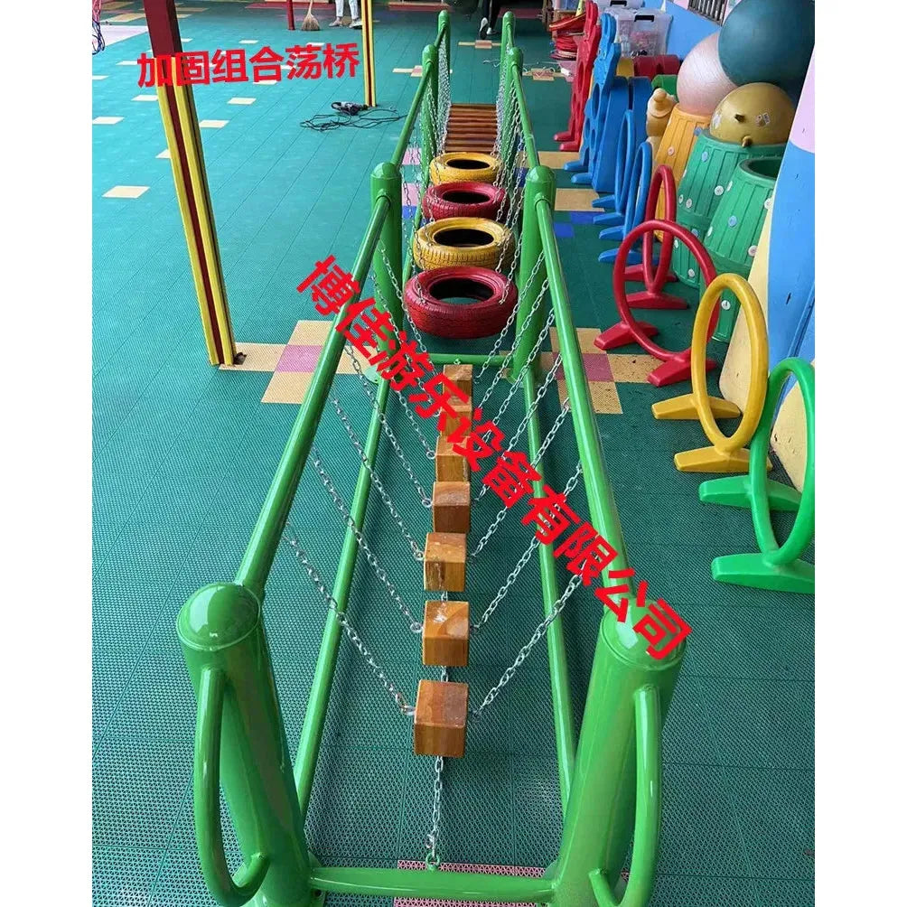 A unisex playground featuring a balance bridge made of wooden blocks and tires linked by chains, suitable for ages 3-8. The area is cushioned with a soft green mat, while colorful rings and a metal toy enhance the playful setting, part of outdoor toys like swings and climbing nets.