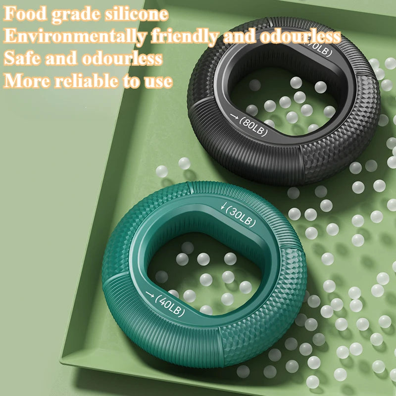 Two circular Silicone Grip Trainer Rings for hand strengthening and wrist rehabilitation, in black and teal, rest on a green surface with white balls scattered around. Text highlights: Food grade silicone, Eco-friendly, Safe and odorless. Ideal for improving grip strength.