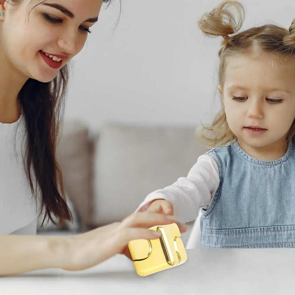 A woman and a young girl with pigtails sit together at a table in Mainland China, smiling as they play with the gold Infinity Gear Cube Spinner Antistress Toy. The woman gently touches the fidget block while the girl watches with interest.